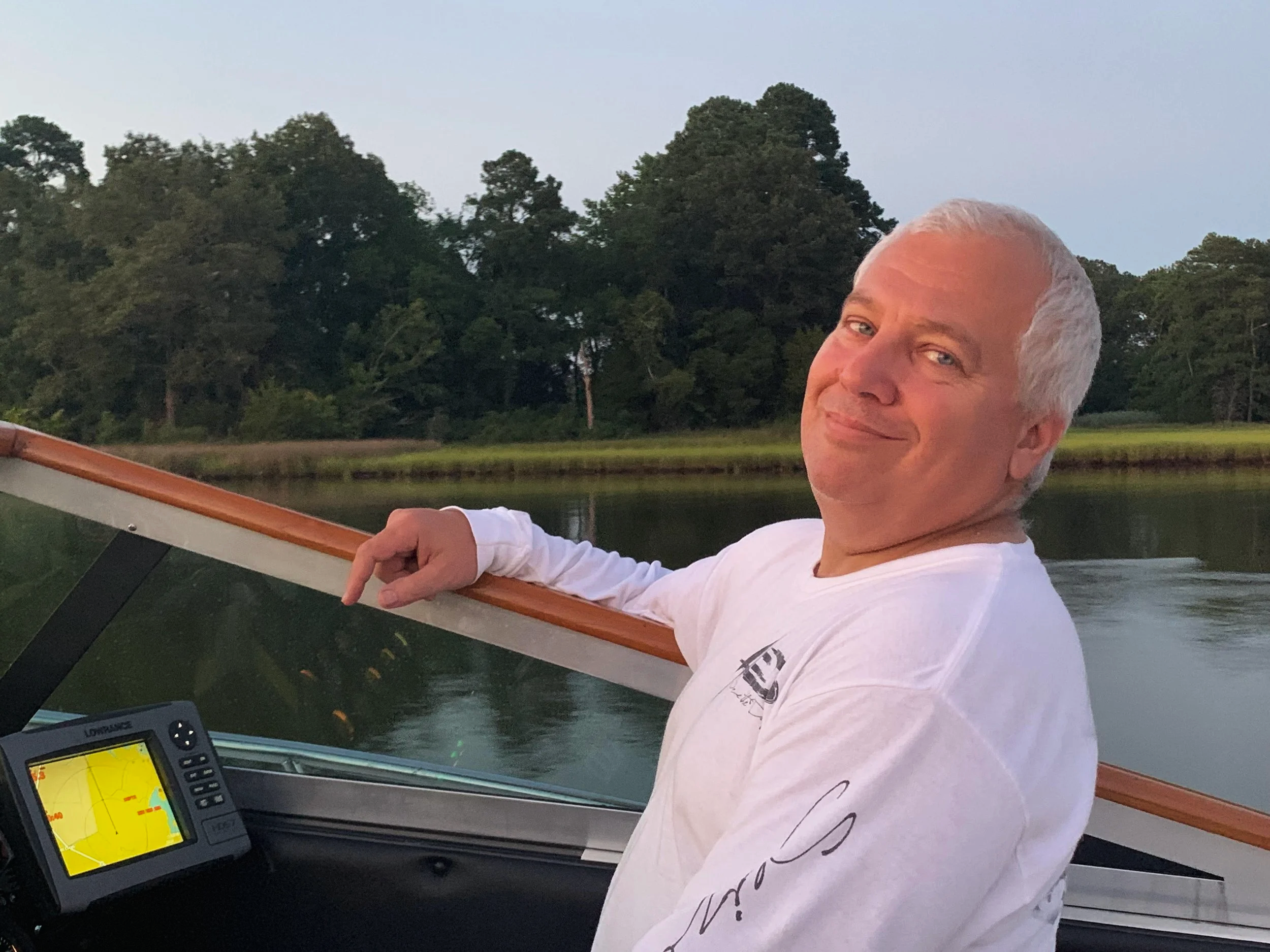 Smiling man with white hair boating on a calm river with lush green trees in the background.