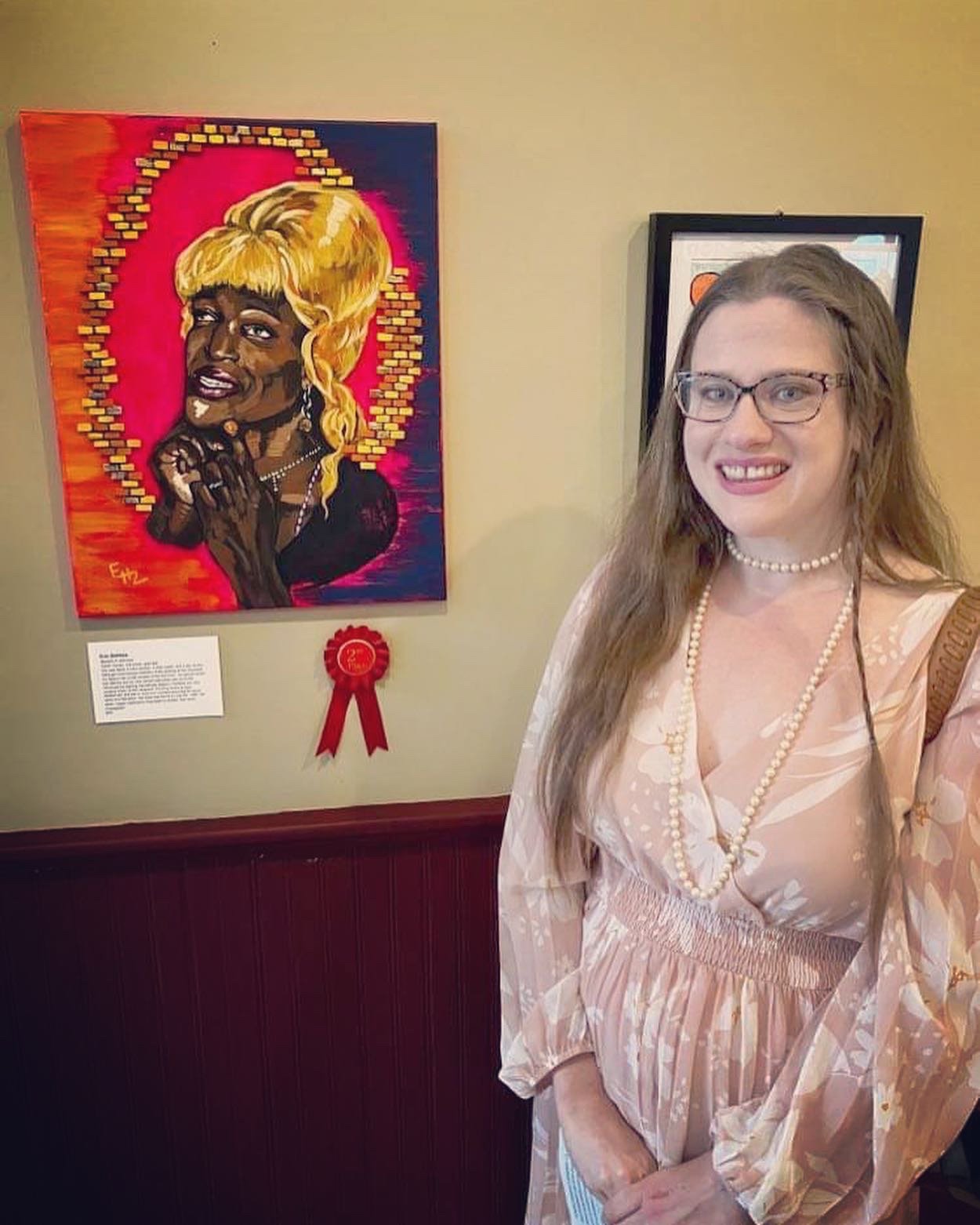 A woman with long brown hair, glasses, wearing a peach-colored floral dress and pearl necklace, standing next to a colorful artwork of a woman with blonde hair, a halo of small square tiles, and a red ribbon award on the wall.