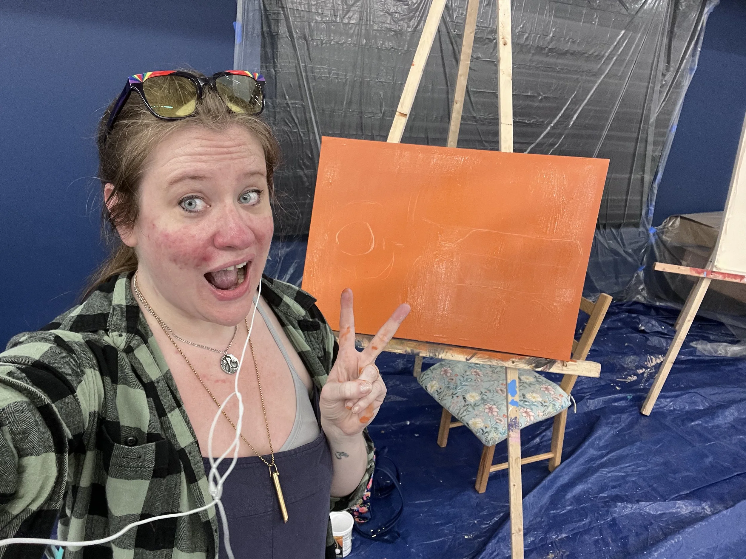 Woman taking a selfie in an art studio, making a peace sign, with orange canvas on an easel behind her, and blue and plastic-covered walls and floor.