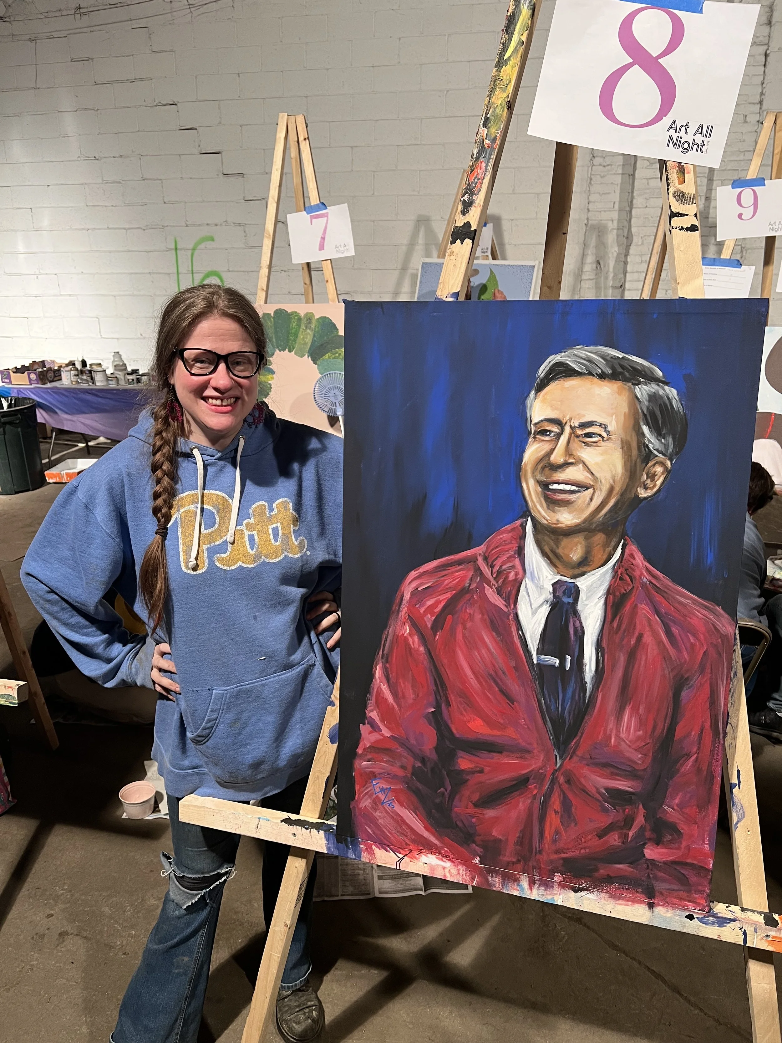 A woman with glasses and a braid, wearing a blue 'Pitt' sweatshirt, smiles and stands next to a large, colorful portrait of a man in a red jacket.