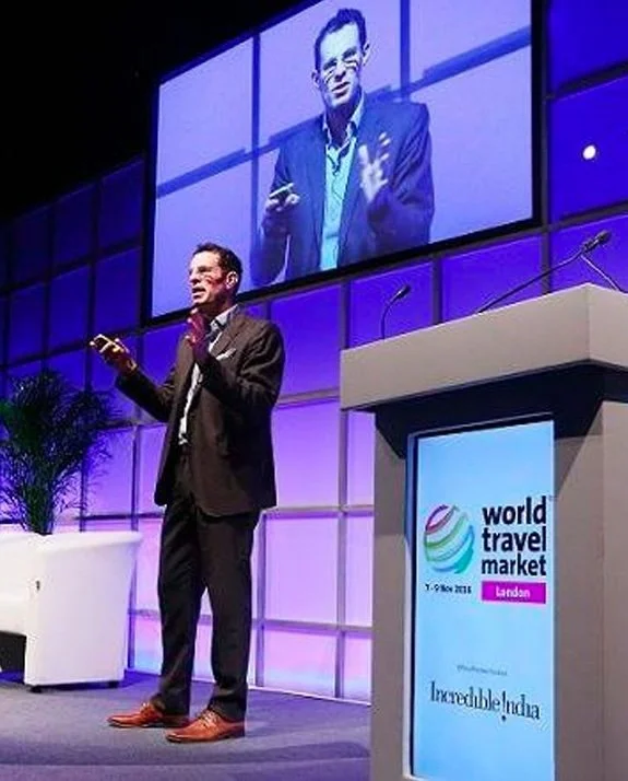 A man in a dark suit giving a presentation at the World Travel Market conference in London, with a large screen behind him displaying his live image, and a podium with the event logo and sponsor information.