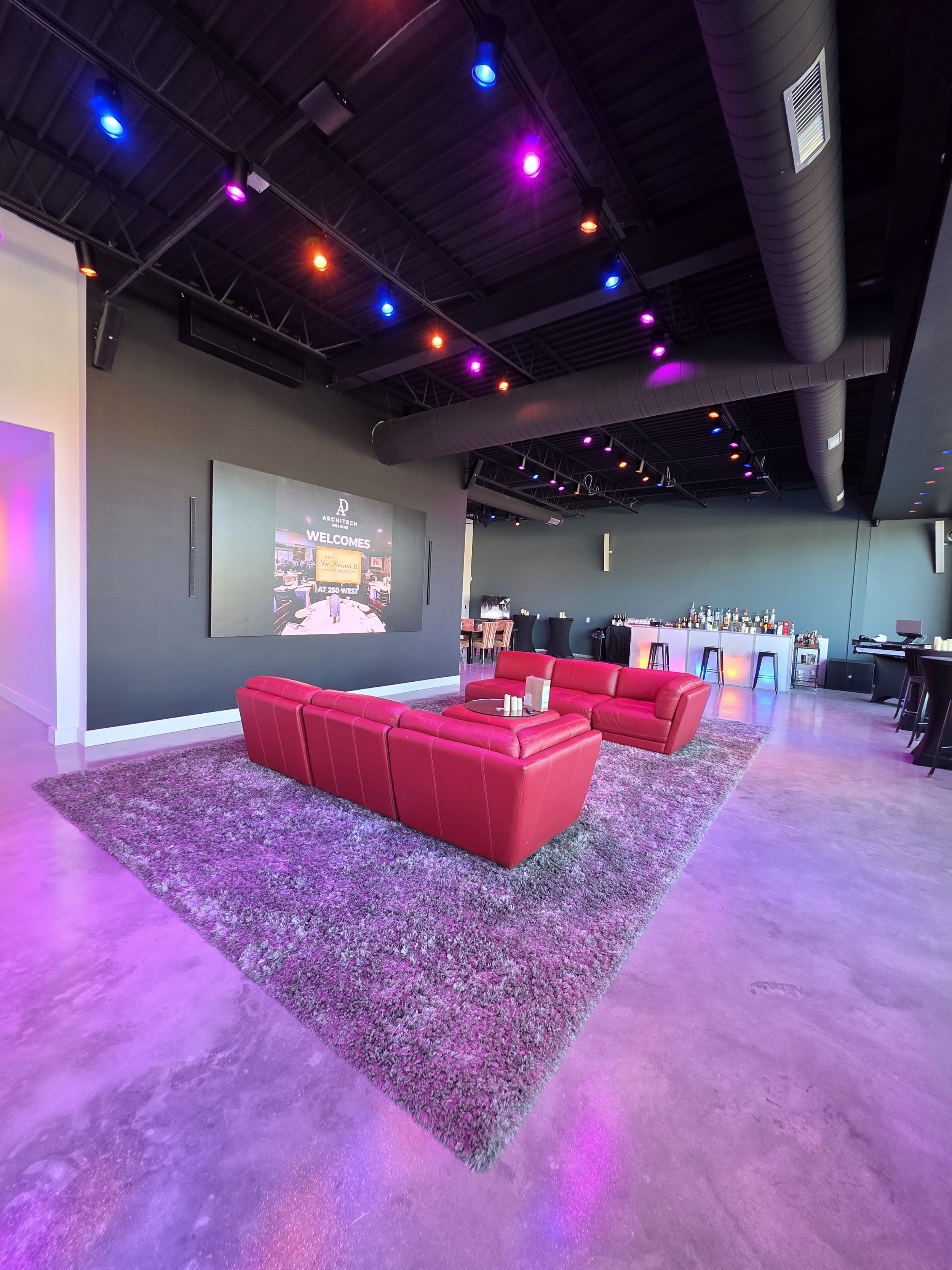 A lounge area with a red sectional sofa on a large, textured gray rug in a modern bar or event space with colorful ceiling lights and a bar area in the background.