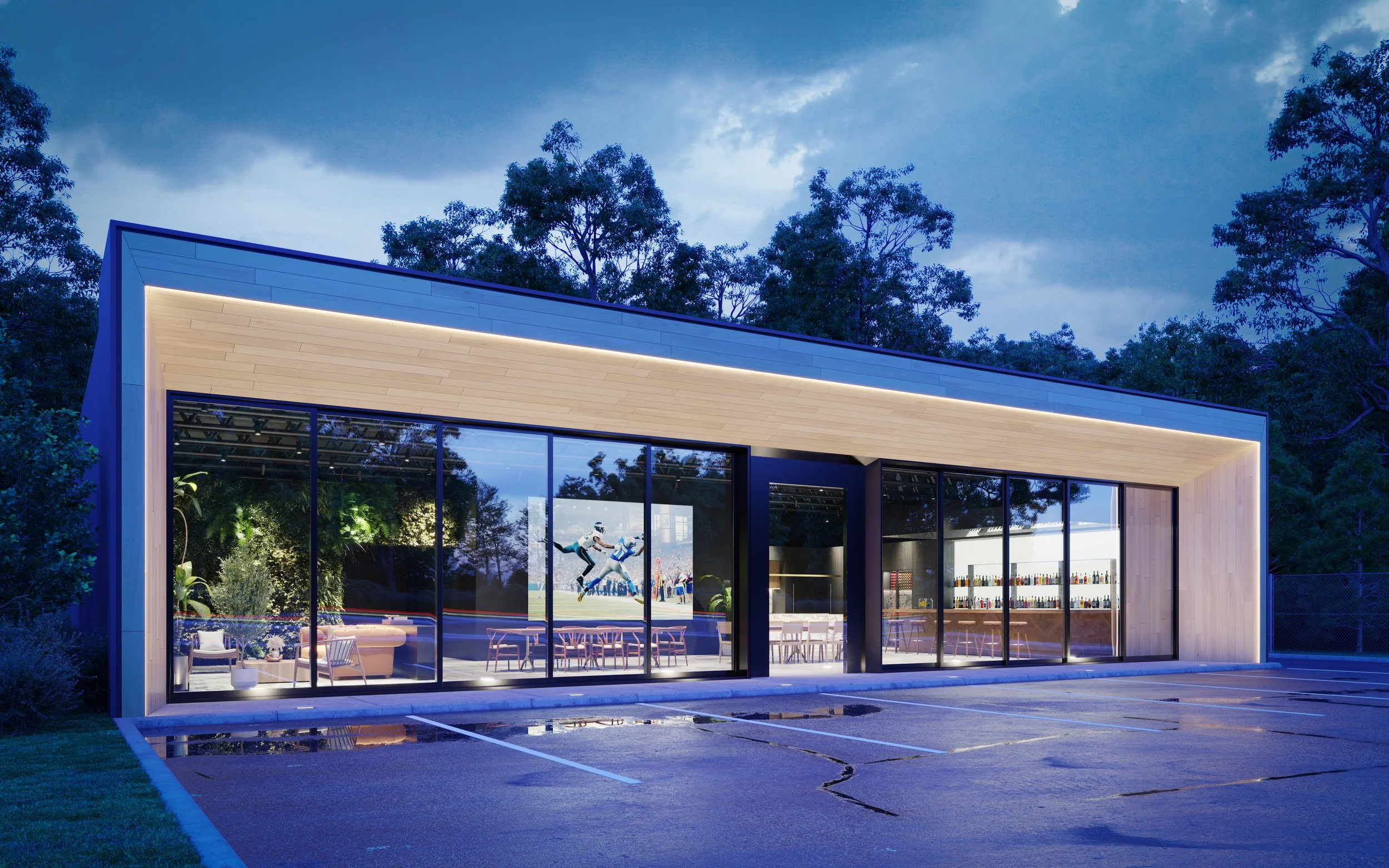 Night view of a modern glass-front building with a parking lot in front, surrounded by trees in the background.