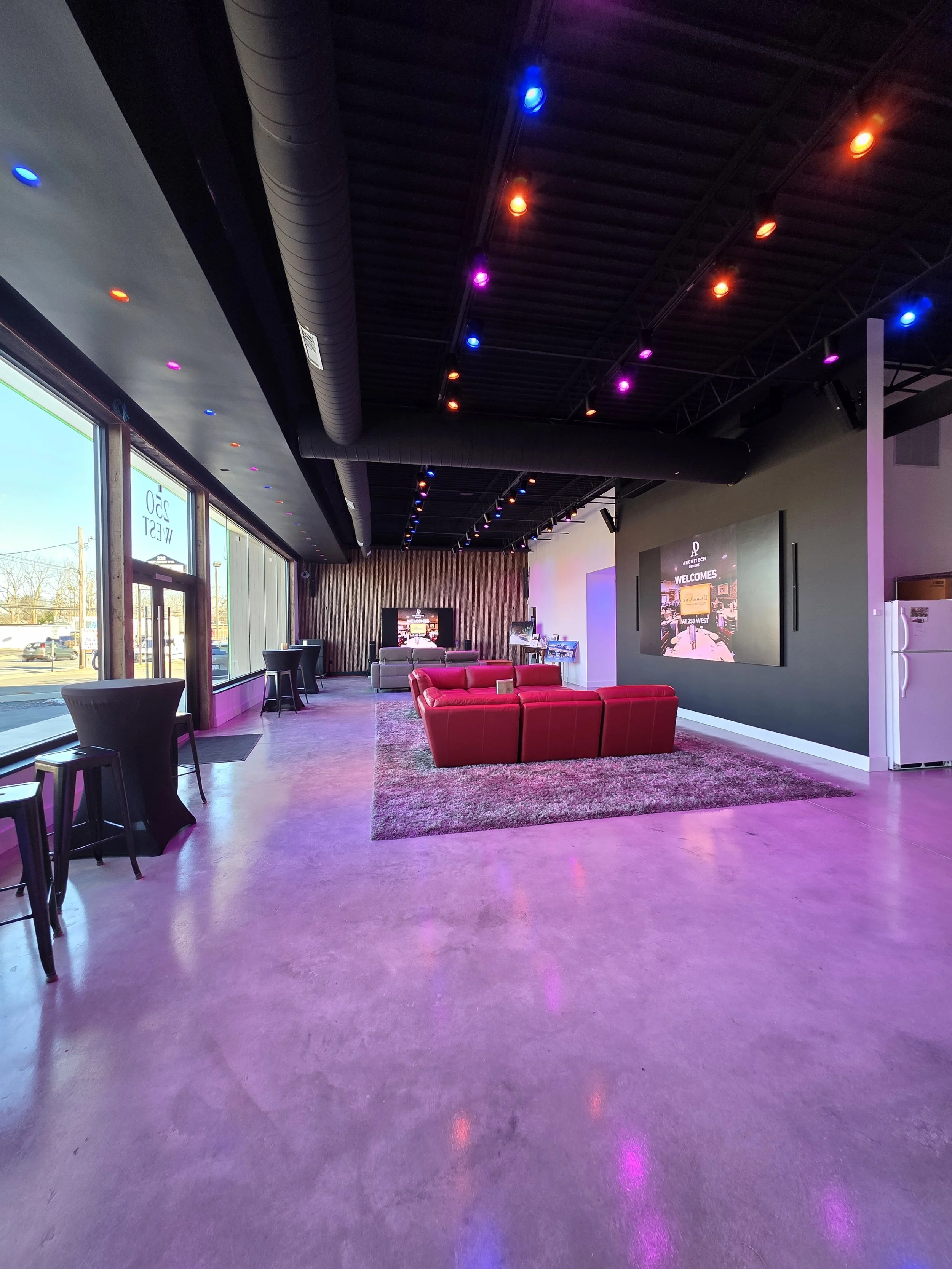 Modern lounge area with red couches, high tables, and a large wall screen, illuminated by colorful ceiling lights.