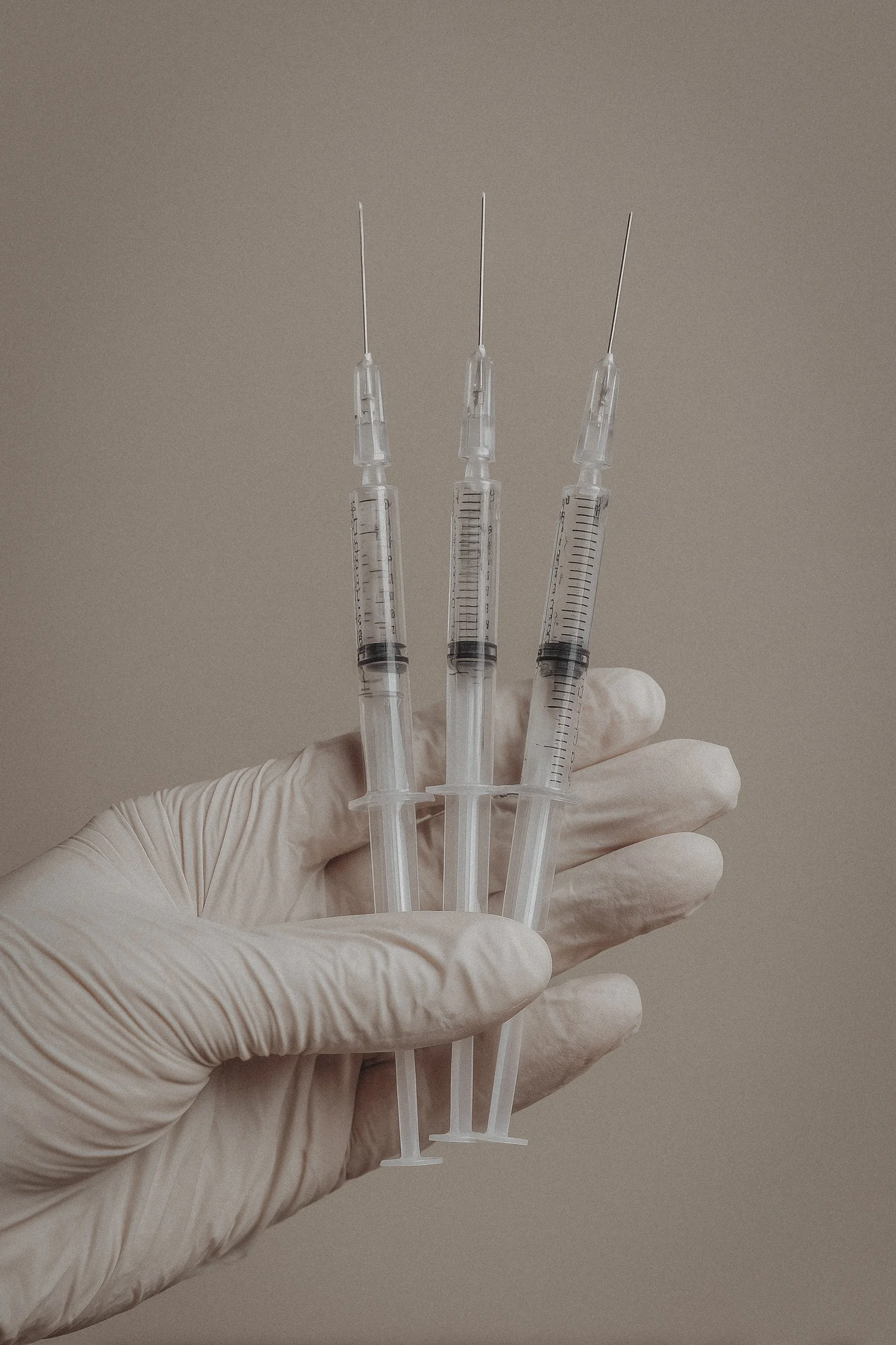 Three syringes held by a hand wearing a latex glove, with a neutral background.