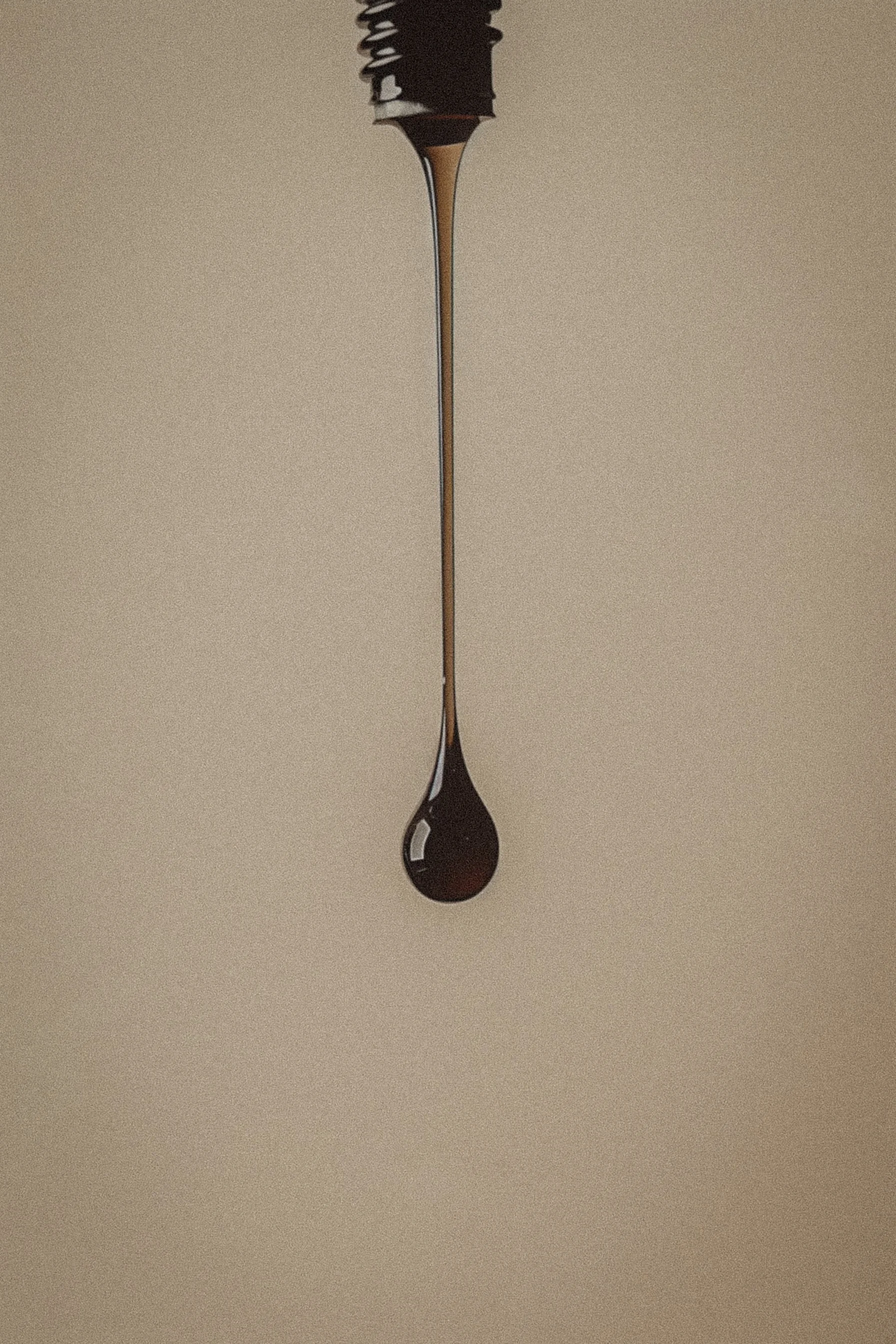 A close-up photograph of a single dark droplet of liquid hanging from a thin stream, against a plain beige background.