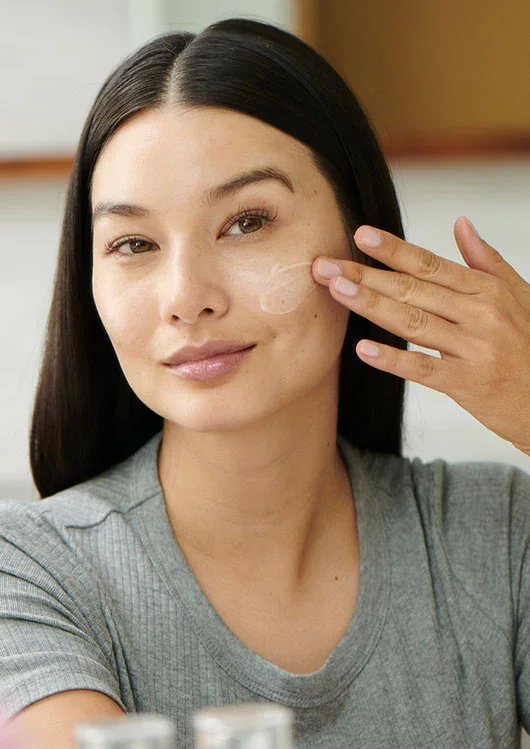 Woman applying facial cream or moisturizer to her cheek in a bathroom or bedroom setting.