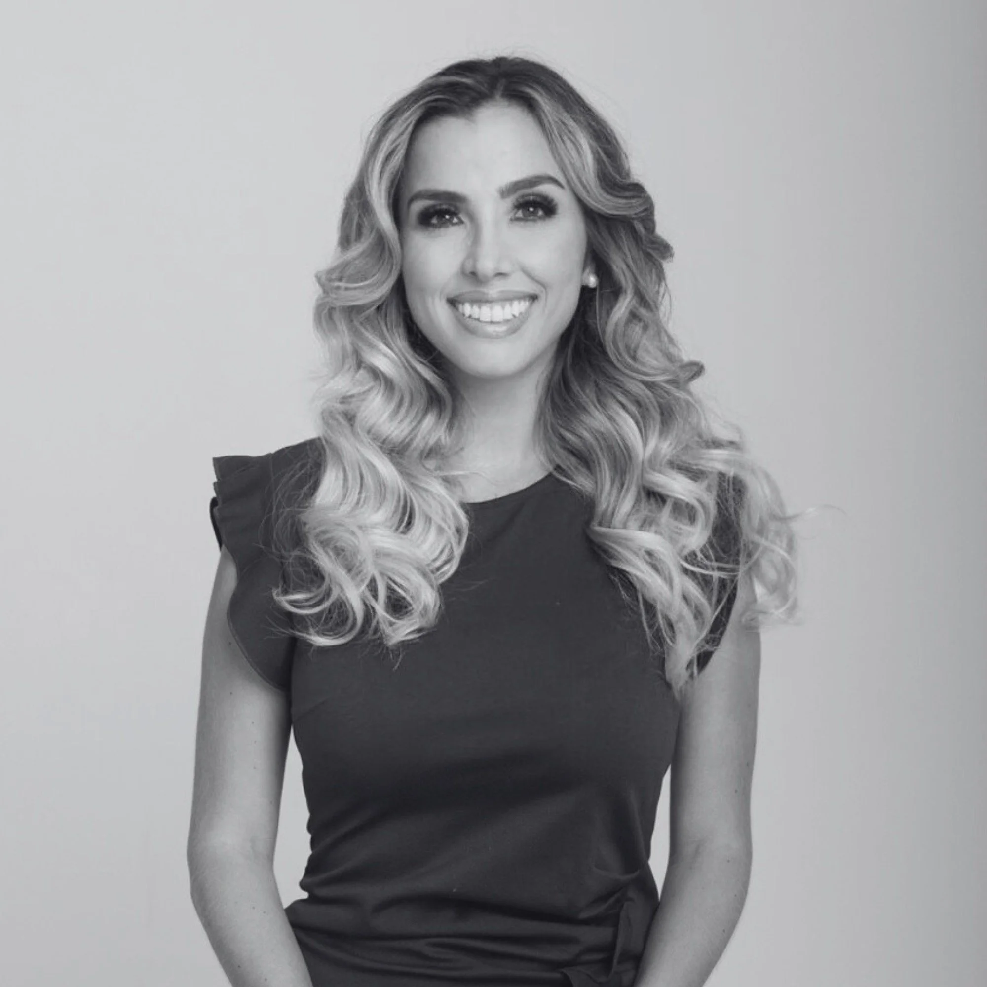 Black and white portrait of a woman with long, wavy hair, smiling, wearing a dark top, standing against a plain light background.