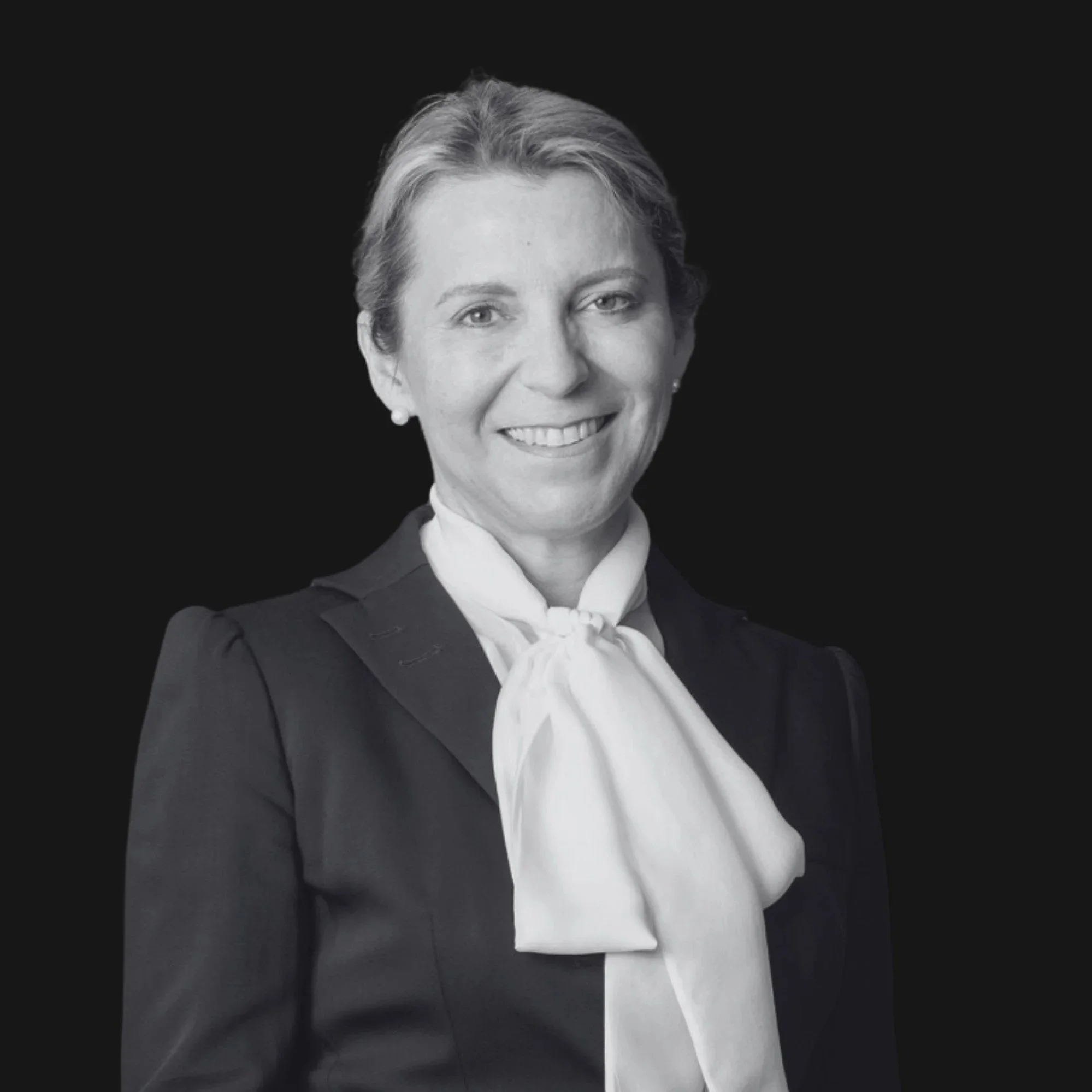 A woman with short gray hair smiling in a black suit and white blouse against a dark background.