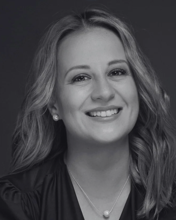 A smiling woman with wavy hair, wearing pearl earrings and a necklace, in a black and white portrait.