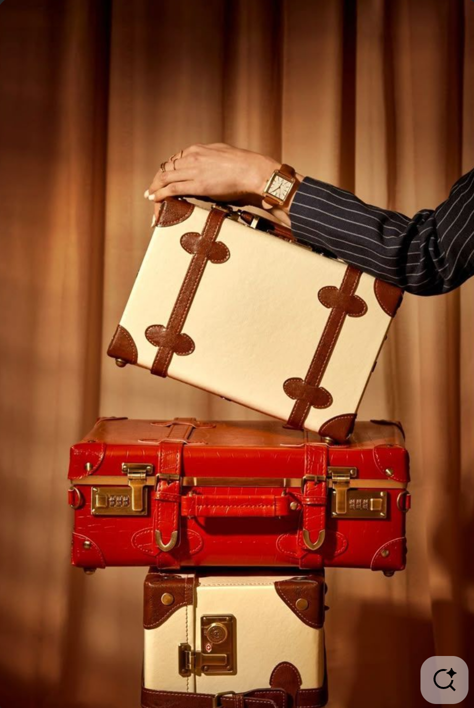 Hand holding a beige and brown patterned suitcase above two stacked suitcases, one red and one white with brown accents, with a woman wearing a striped blazer and a gold watch in the background.