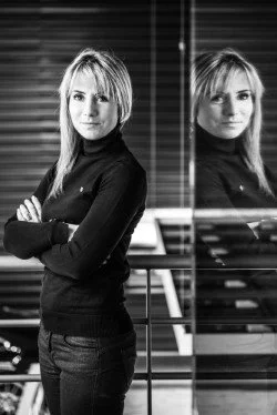 A woman with shoulder-length blonde hair wearing a black turtleneck sweater standing next to a glass wall with reflections.