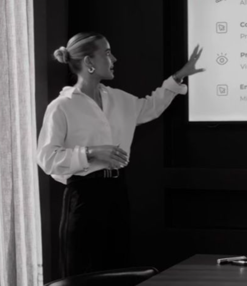 A woman in a white blouse presenting in front of a large screen with a presentation slide, gesturing with her right hand, in a professional setting.