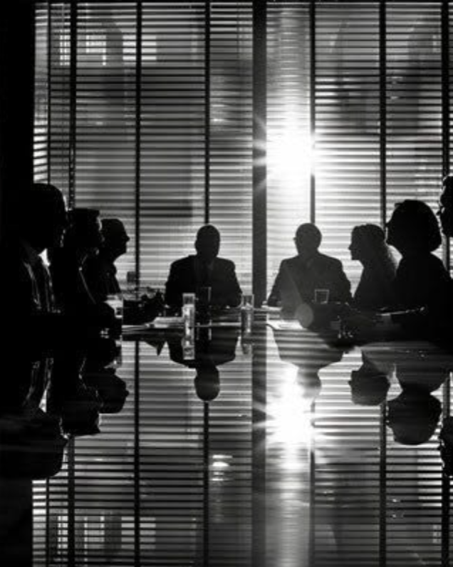 Silhouettes of business people sitting around a conference table in a meeting room with window blinds, sunlight shining through, and their reflections on the shiny table surface.