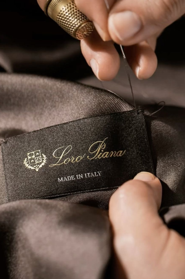 Close-up of a person sewing a clothing label that reads "Loro Piana, Made in Italy" with a needle and thread.