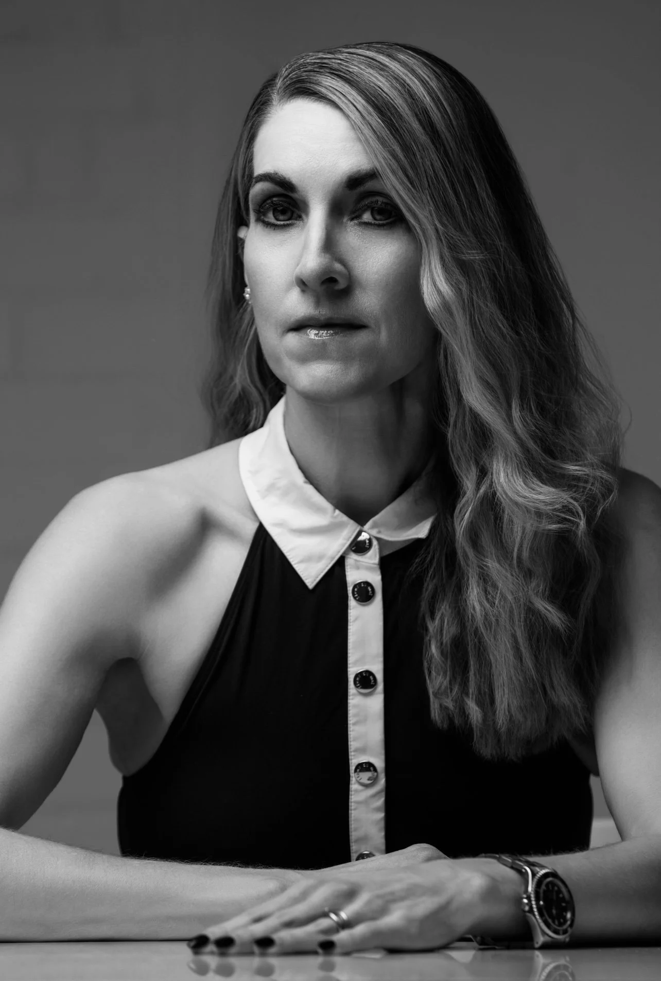A black and white portrait of a woman with long wavy hair, wearing a sleeveless high-neck blouse with buttons, sitting at a table with her arms resting on it, and a watch on her wrist.