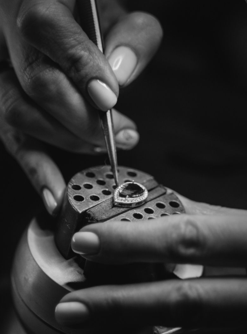Hands holding jewelry and a tool, working on a jewelry setting in a workshop.