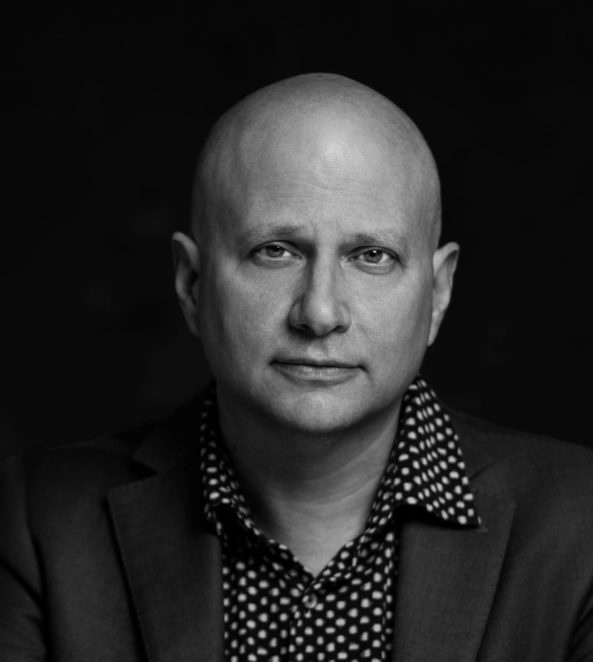 Close-up black and white portrait of a man with a completely shaved or bald head, wearing a polka-dot shirt and a blazer.