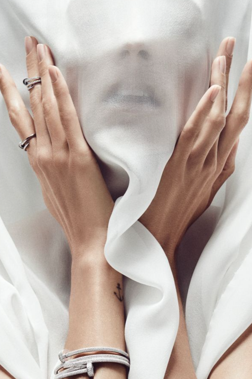 Two hands with rings and bracelets are holding a semi-transparent white fabric that reveals a person's face behind it.