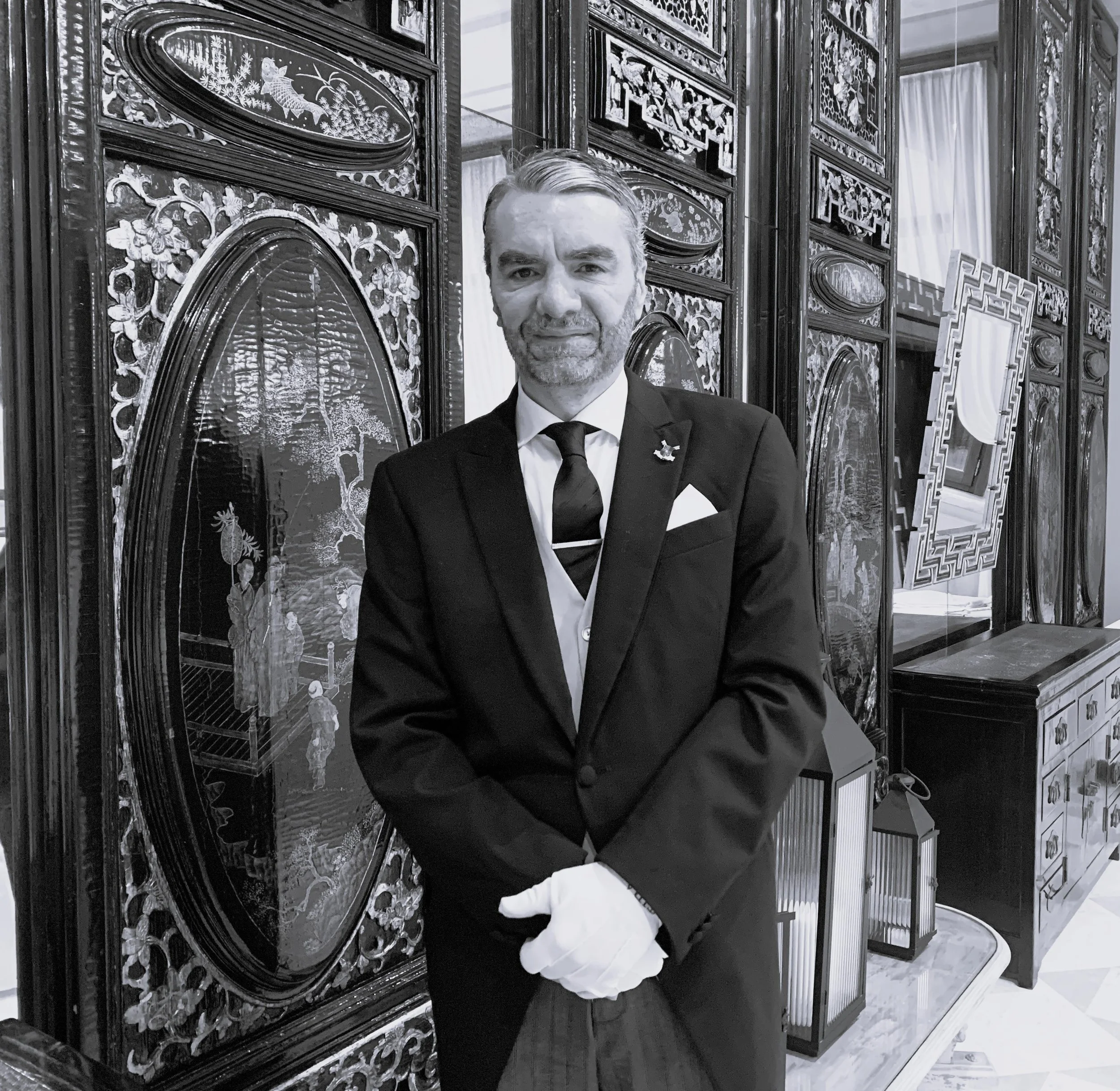 A man in a suit and tie standing in front of ornate decorative screens with floral and nature patterns, in a luxurious or formal setting.
