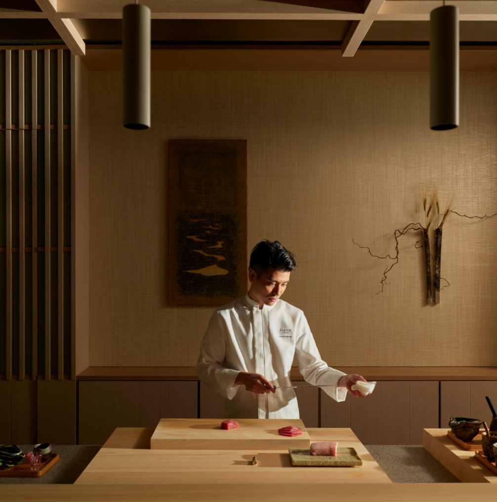 A chef in a white uniform preparing slices of sashimi at a wooden counter in a modern, minimalist restaurant with Japanese decor.