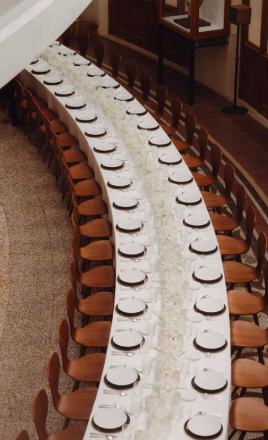 A long banquet table set with white tablecloths, black plates, and glassware, arranged in a curved formation in an elegant room.