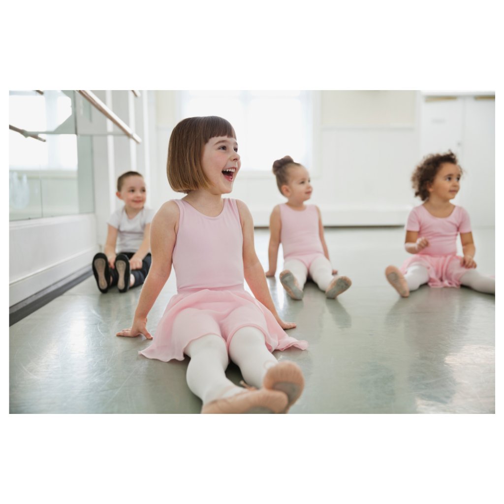 Four young girls sitting and smiling in a ballet studio, wearing pink and white ballet outfits.