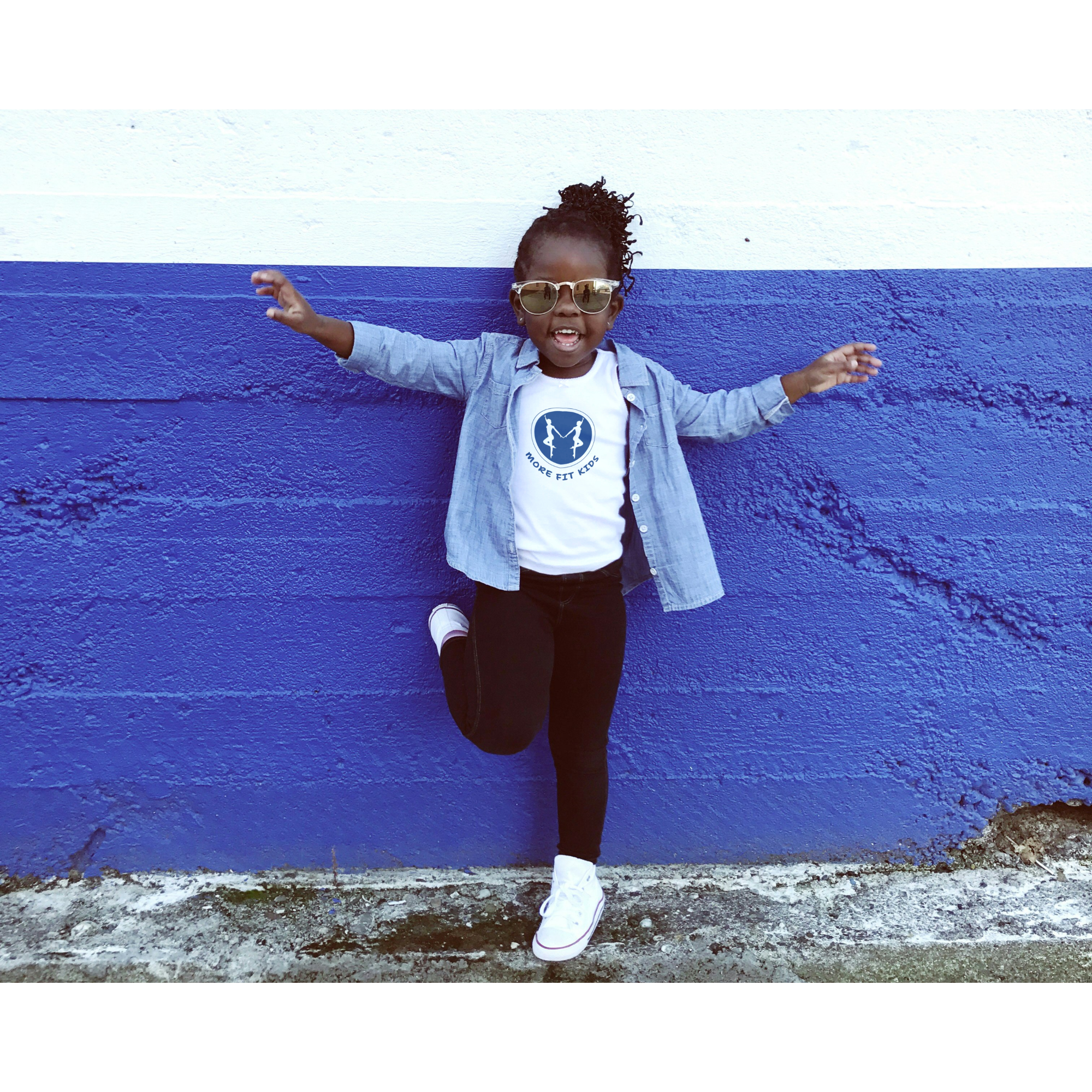 Young girl with curly hair, sunglasses, white sneakers, gray shirt, white t-shirt with 'More Fit Kids' logo, standing in front of a blue and white painted concrete wall with outstretched arms, smiling.