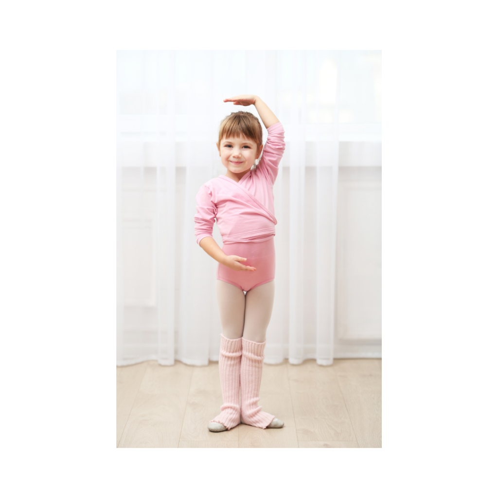 A young girl dressed in pink ballet attire, standing on a wooden floor in front of white curtains, smiling, with her right arm raised above her head and left hand on her waist.