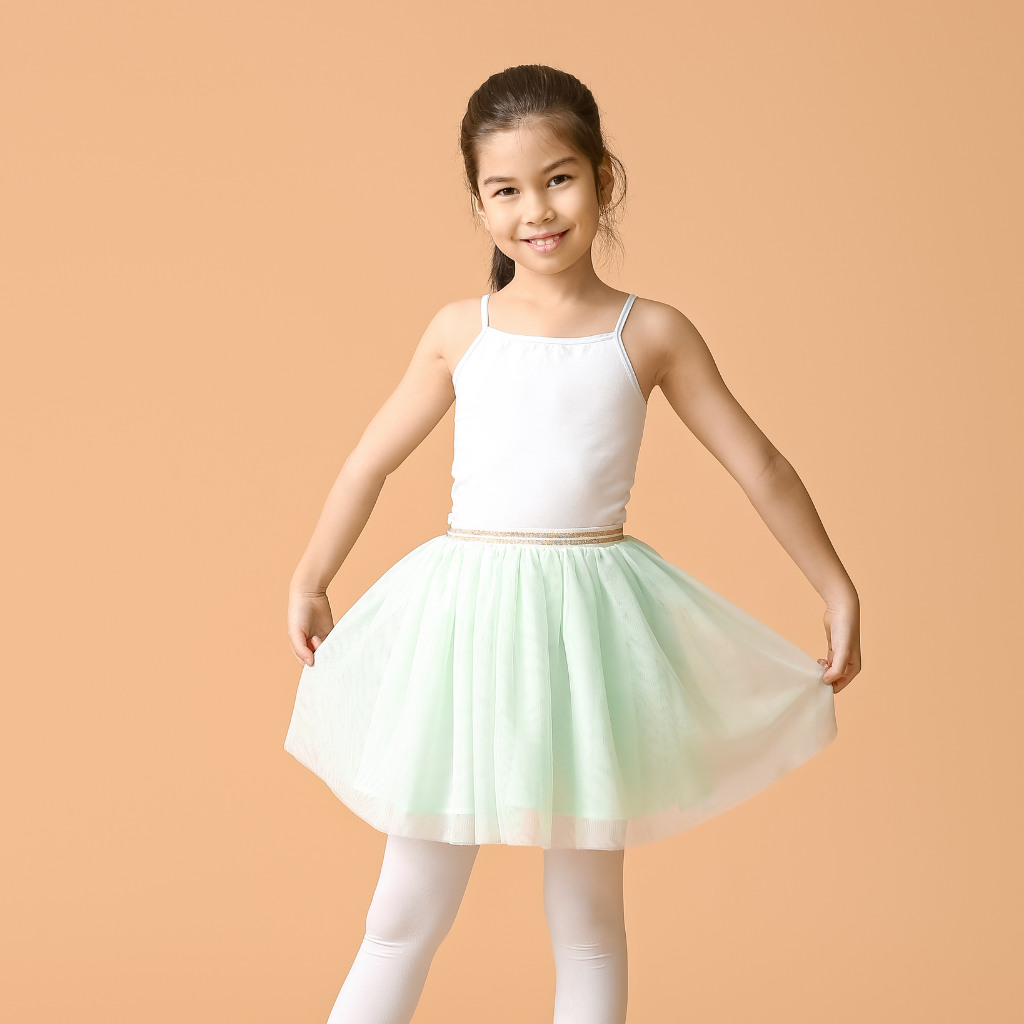 A young girl with dark hair in a ponytail, smiling, dressed in a white tank top, a light green tulle skirt, and white tights, standing against a peach-colored background.
