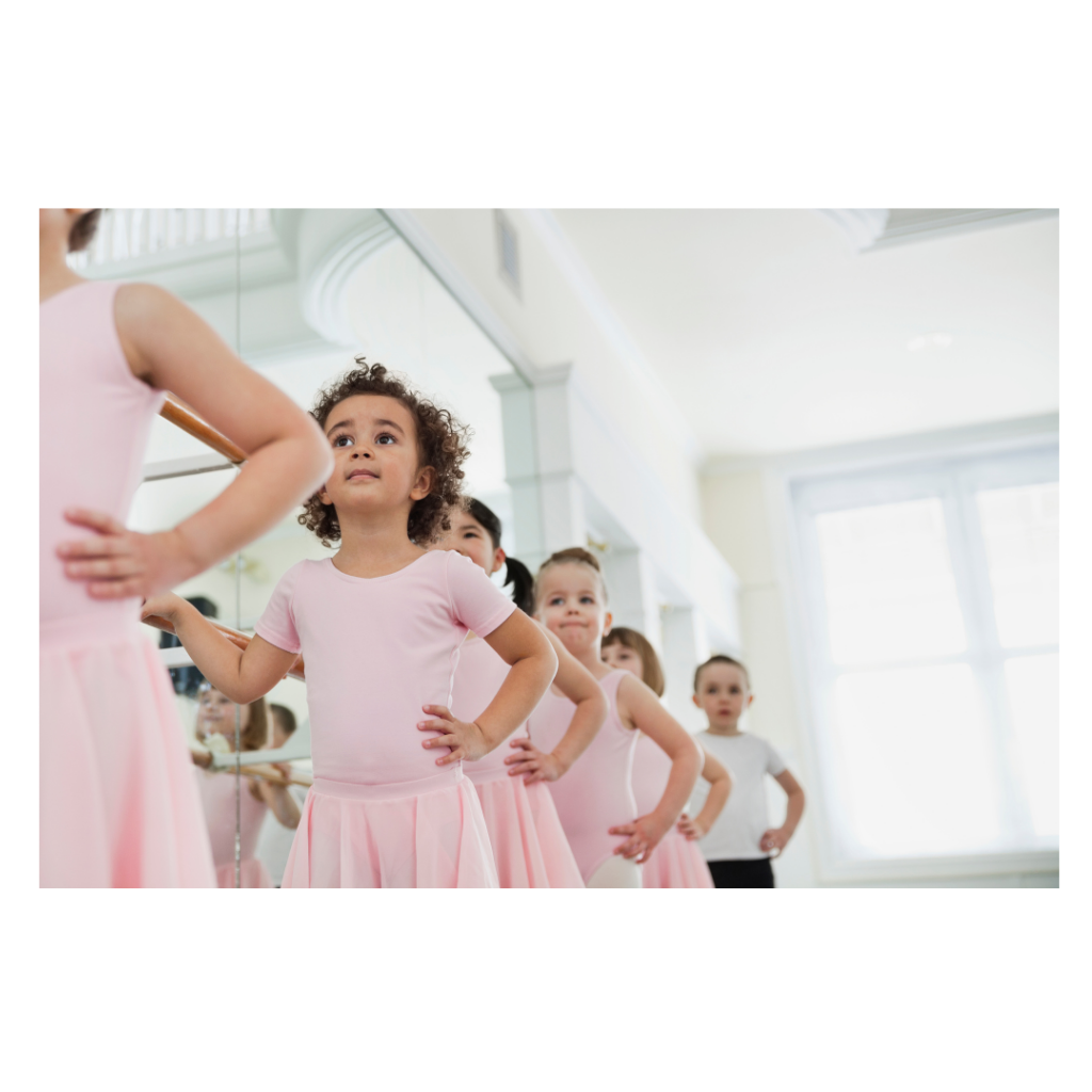 Young girls in pink ballet leotards and skirts standing in front of a mirror in a dance studio, preparing for a dance class.