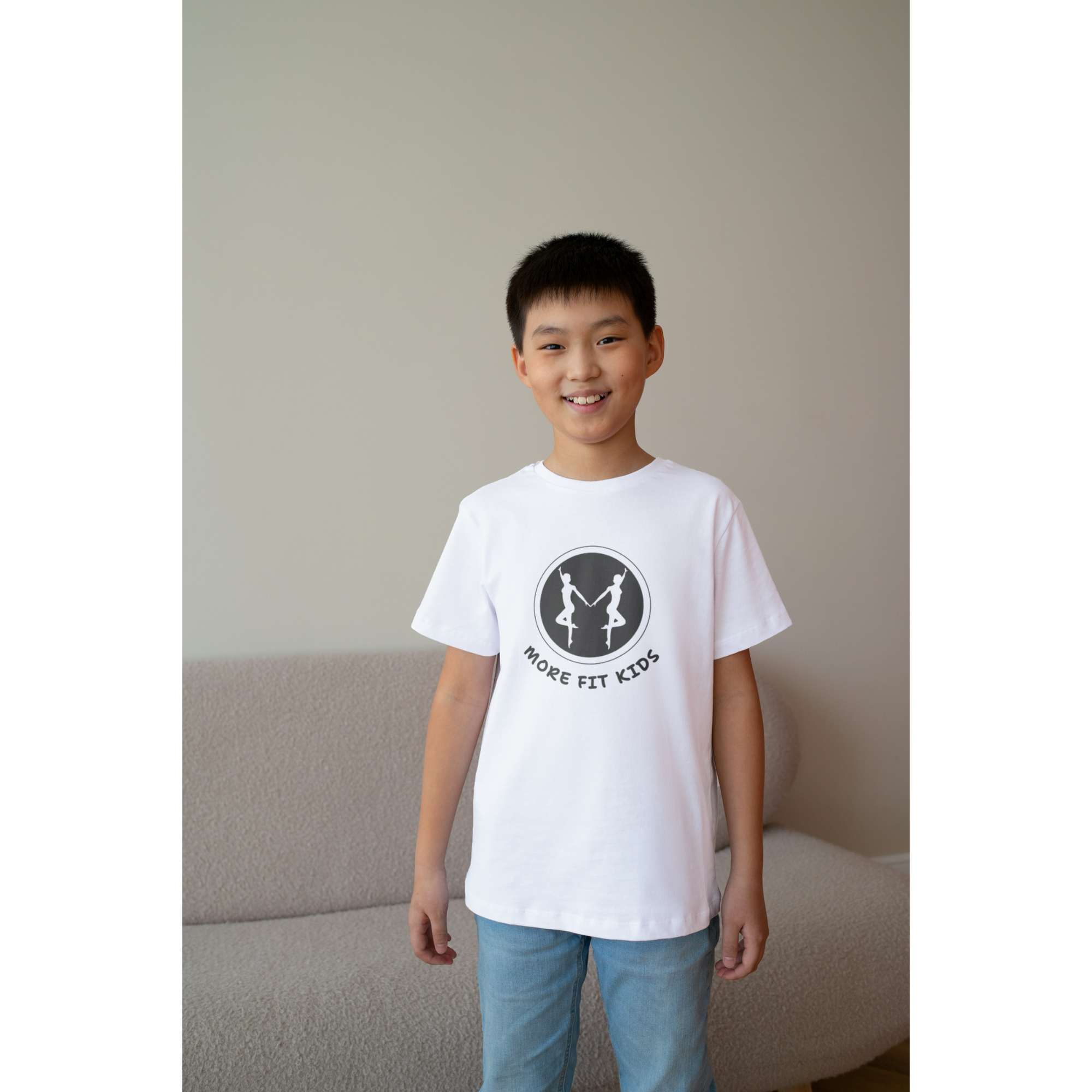 A young boy standing indoors, smiling, wearing a white t-shirt with a logo and blue jeans.