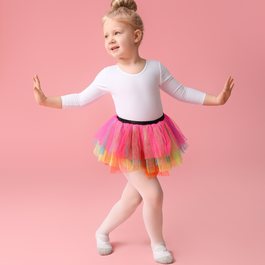 Young girl stretching or dancing, wearing a white long sleeve top, colorful tutu skirt, white tights, and gray shoes, standing against a pink background.