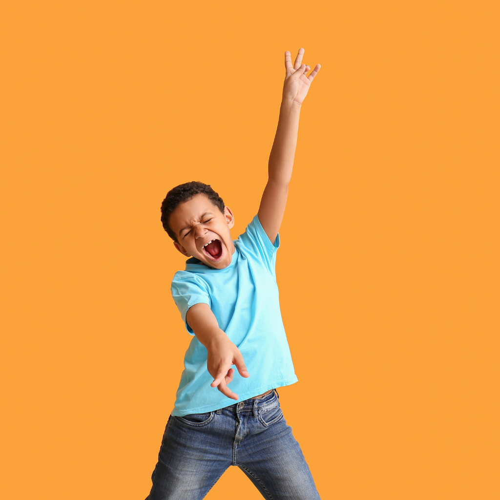 A young boy wearing a blue t-shirt and jeans is joyfully dancing or celebrating with his eyes closed, mouth open, and one arm raised high against an orange background.