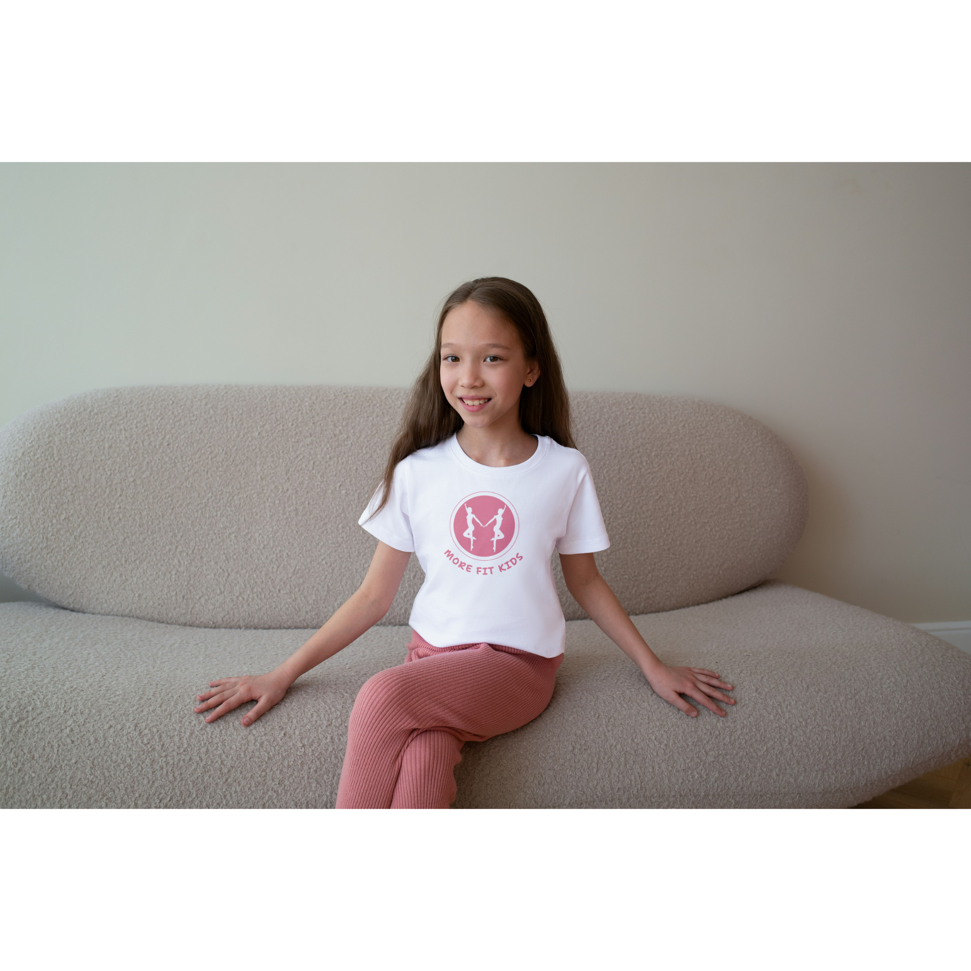 A young girl with long brown hair, smiling, sitting on a beige couch and wearing a white t-shirt with a pink logo and pink pants.