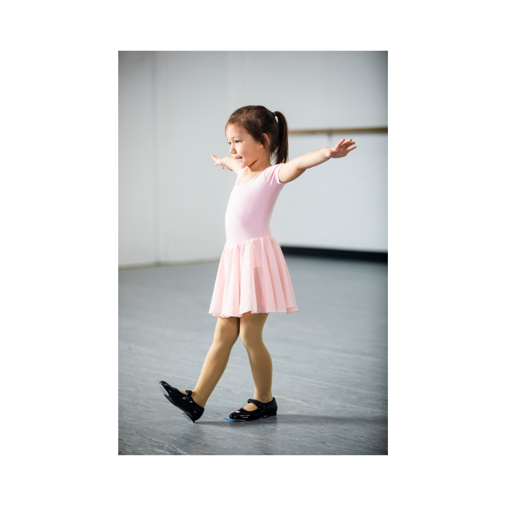 A young girl in a pink ballet dress practicing ballet in a dance studio, with her arms outstretched and a smile on her face.