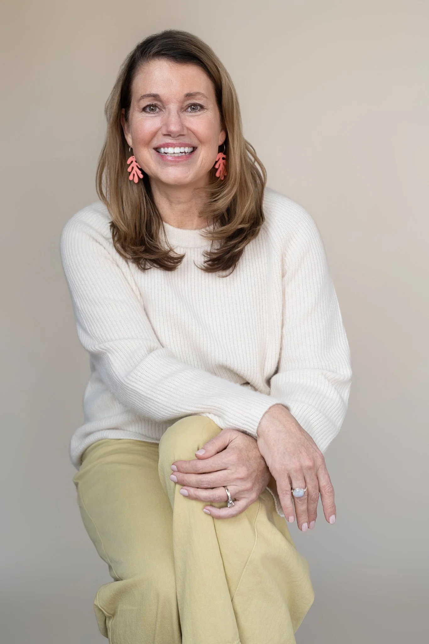 Woman smiling, wearing pink leaf-shaped earrings, a white sweater, and beige pants, sitting against a plain light-colored background.