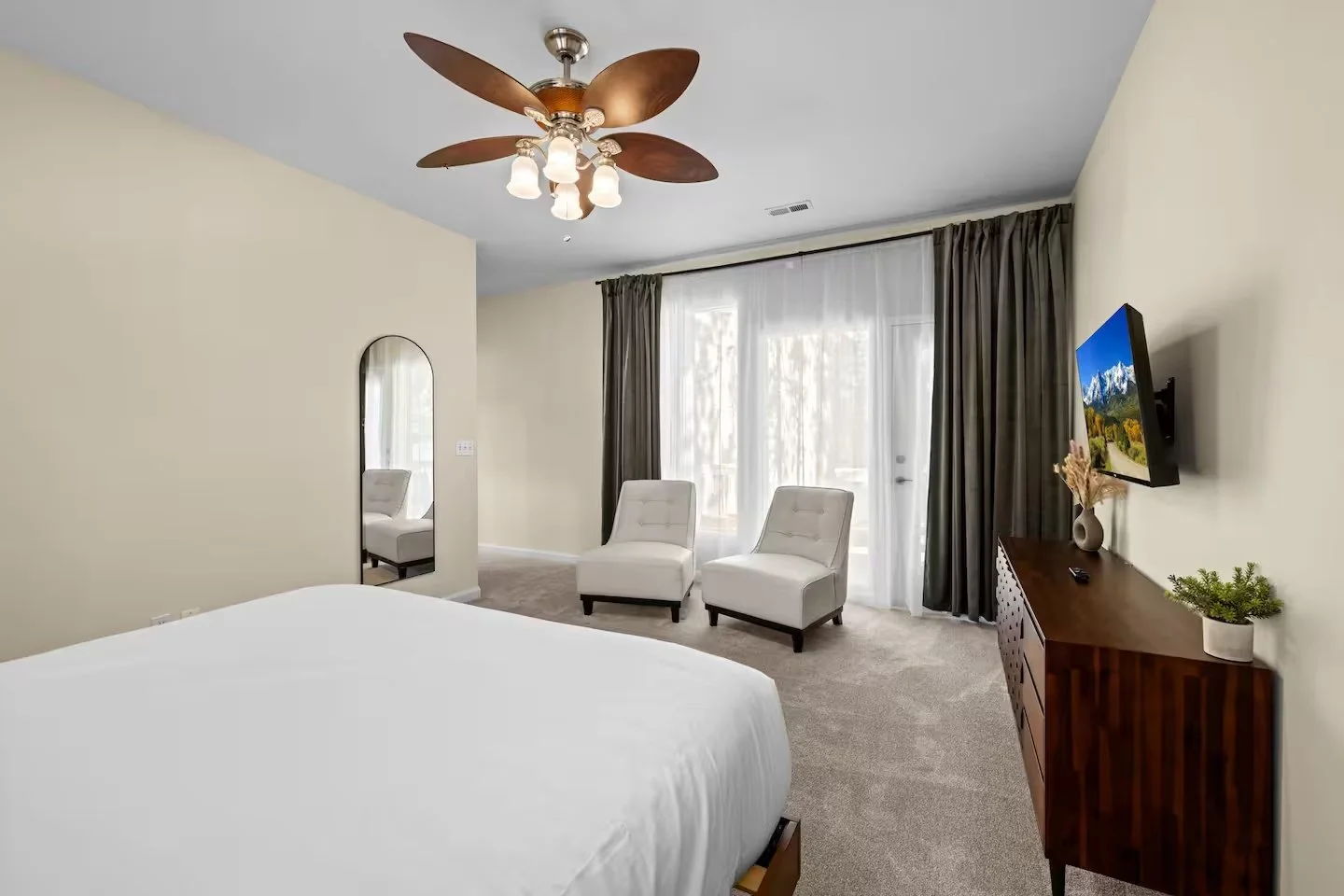 Hotel bedroom with a large bed, two white chairs near a sliding glass door with curtains, a wooden dresser with a TV on the wall, and a ceiling fan with wooden blades.