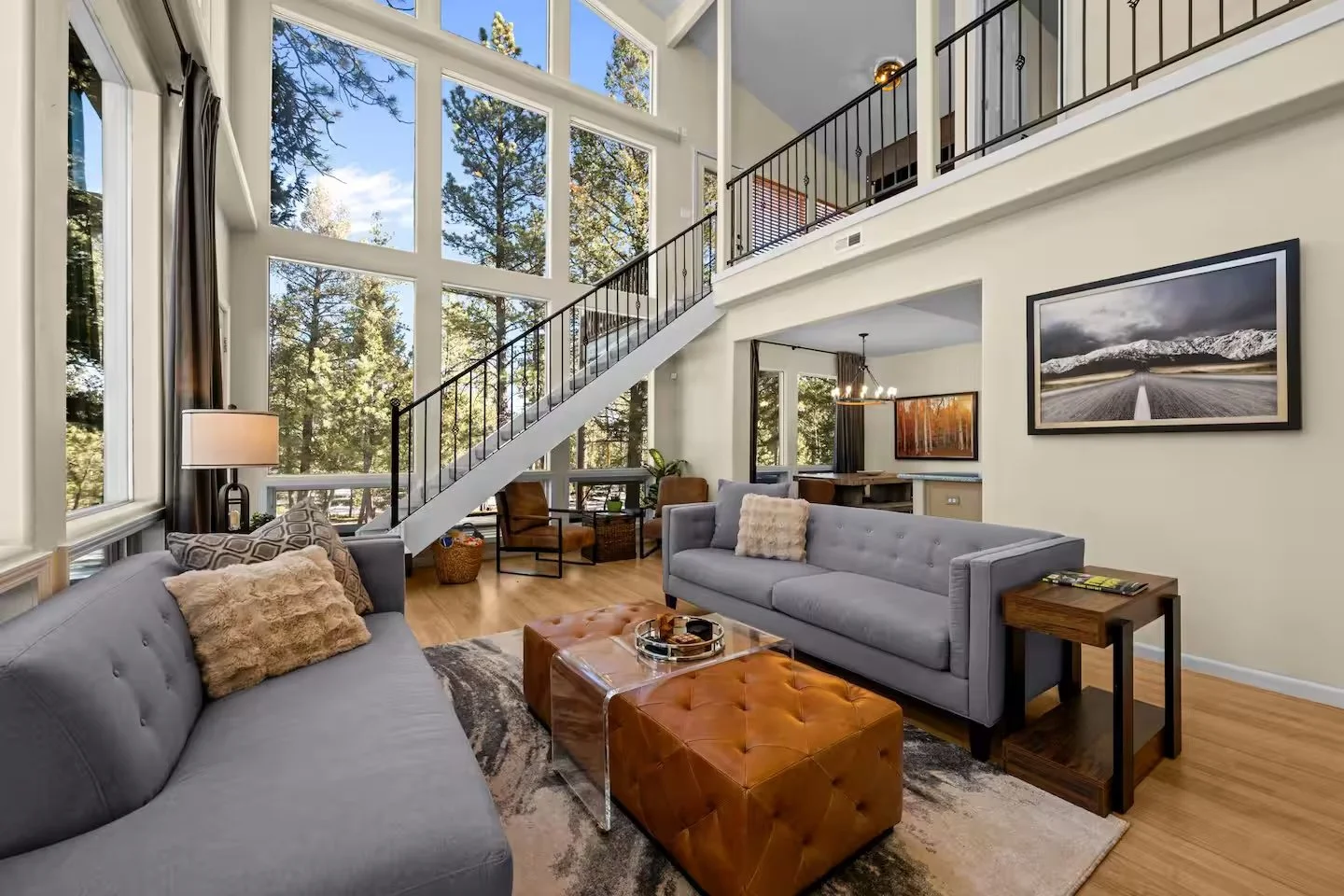 Interior of a modern living room with large floor-to-ceiling windows showcasing a forest view, furnished with sofas, a leather ottoman, side tables, and wall art.