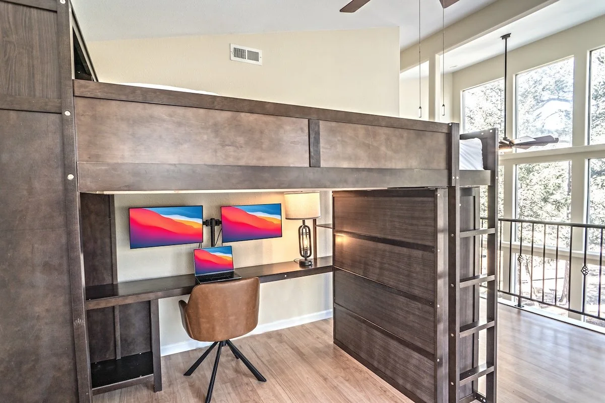 A loft bed with a workspace underneath, including two monitors, a laptop, a desk lamp, and a brown chair, in a bright room with large windows.