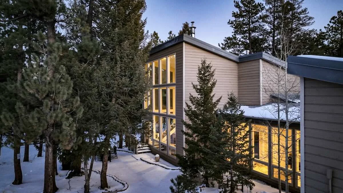 Modern house with large windows illuminated from inside, surrounded by snow and pine trees, in a wooded area during dusk.