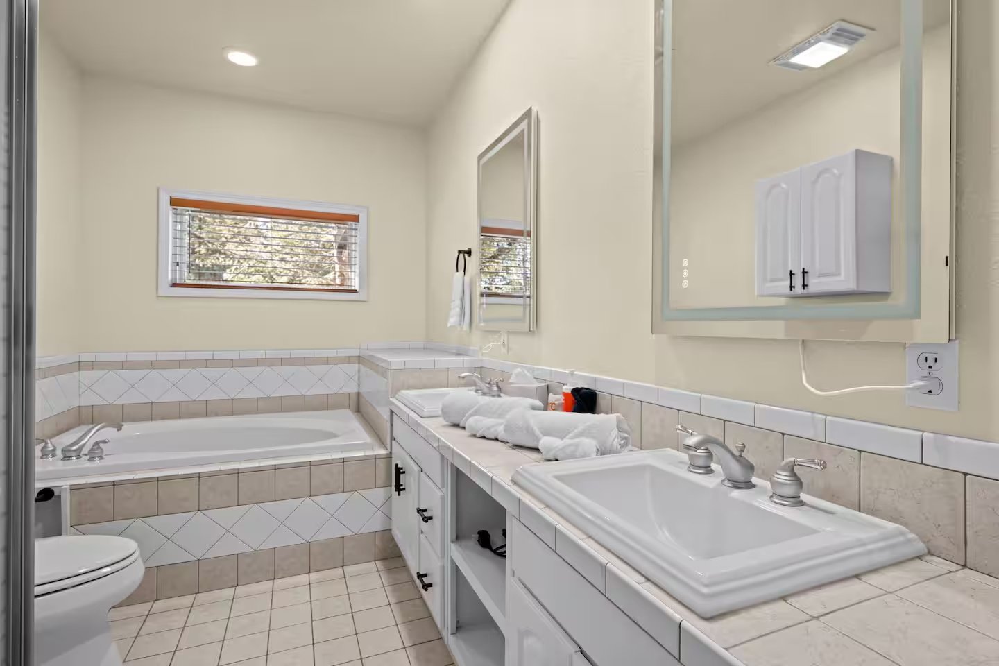 Bathroom with bathtub, double sinks, mirror, window, and tiled surfaces.
