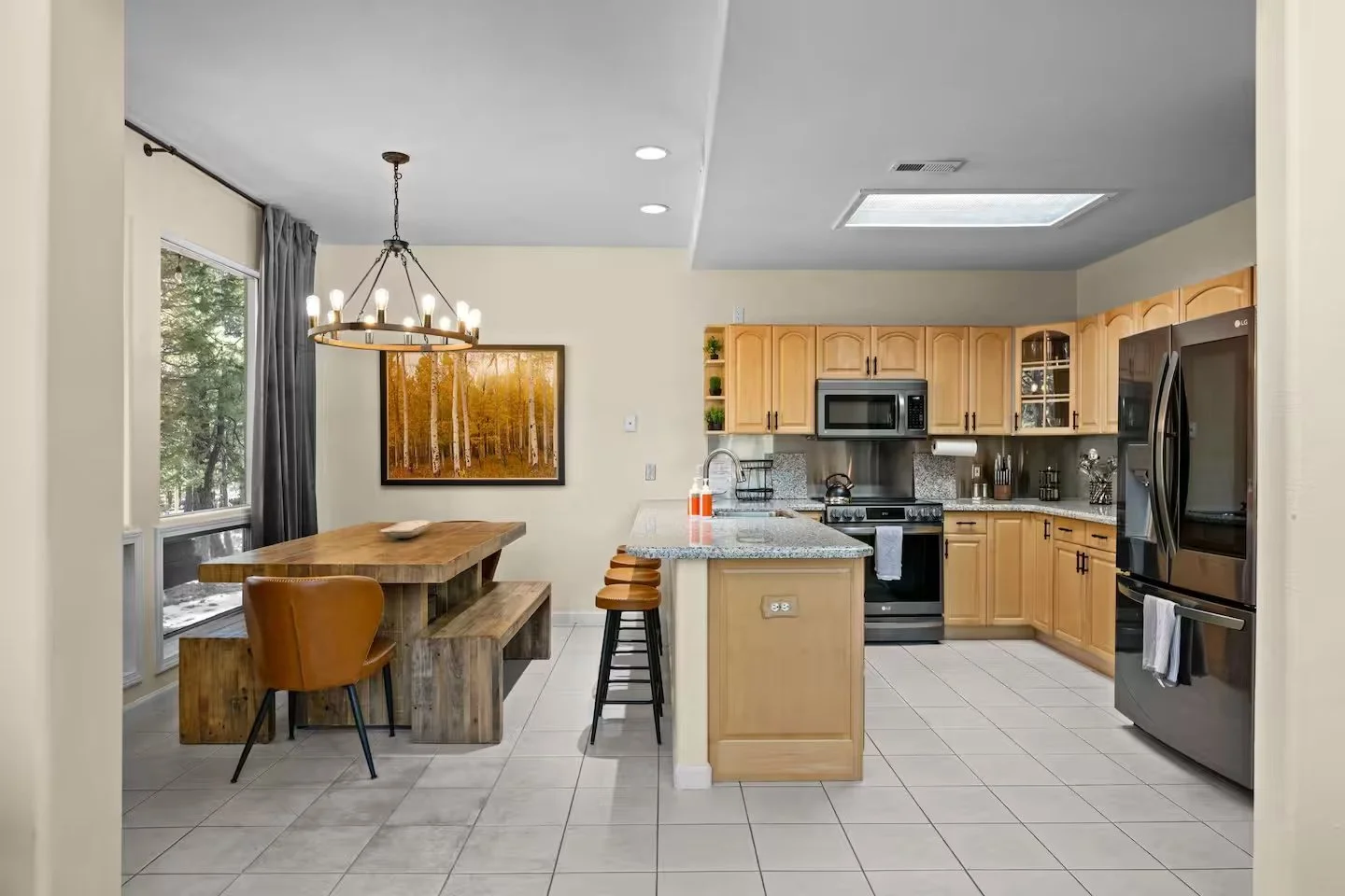 Open-concept kitchen and dining area with wooden dining table, bar stools, light wood cabinets, stainless steel appliances, window with curtains, modern lighting, and artwork on wall.