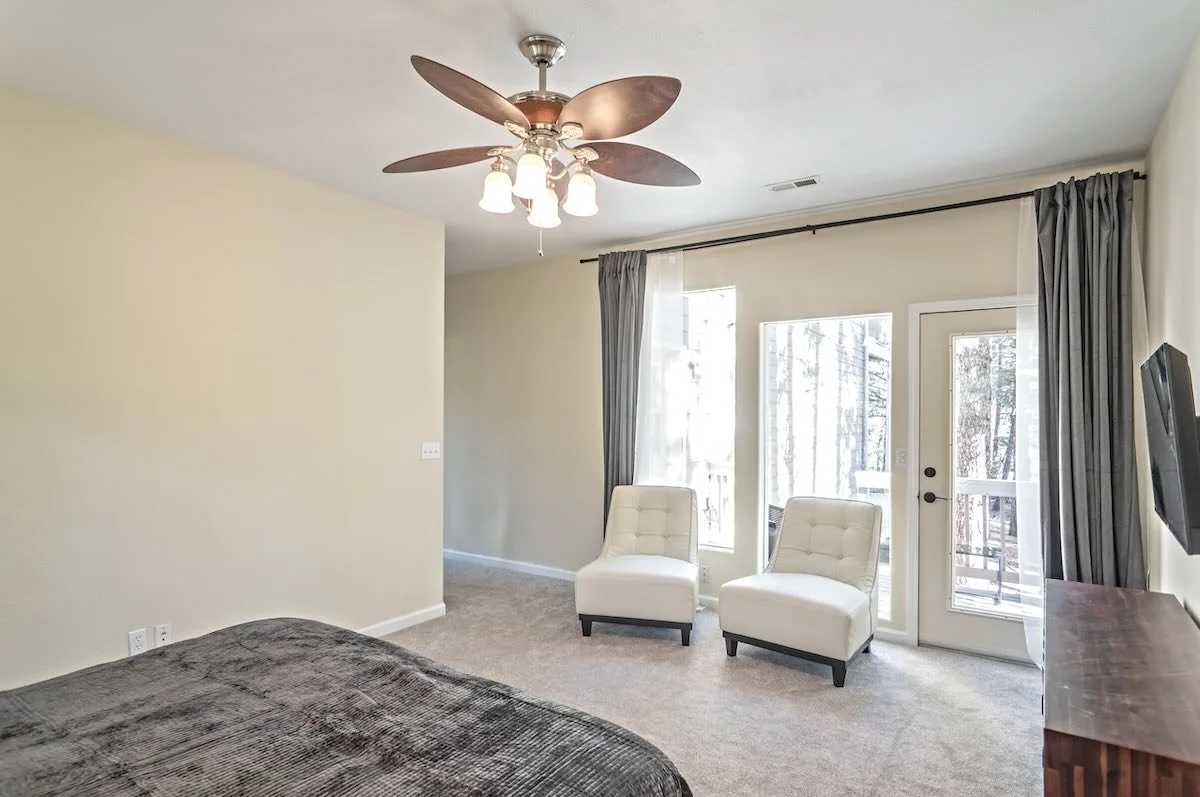 A bedroom with a ceiling fan, two white chairs near glass doors and windows, a brown dresser, and a wall-mounted TV.