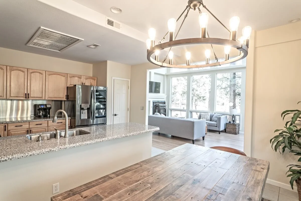 Open-concept kitchen and living room with large window, chandelier, wooden dining table, granite countertops, stainless steel appliances, and light-colored furniture.