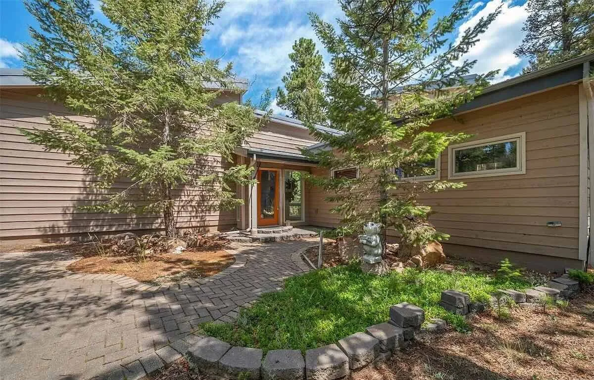View of a house entrance with a small brick patio, surrounded by green trees and a garden area with grass and decorative stones.