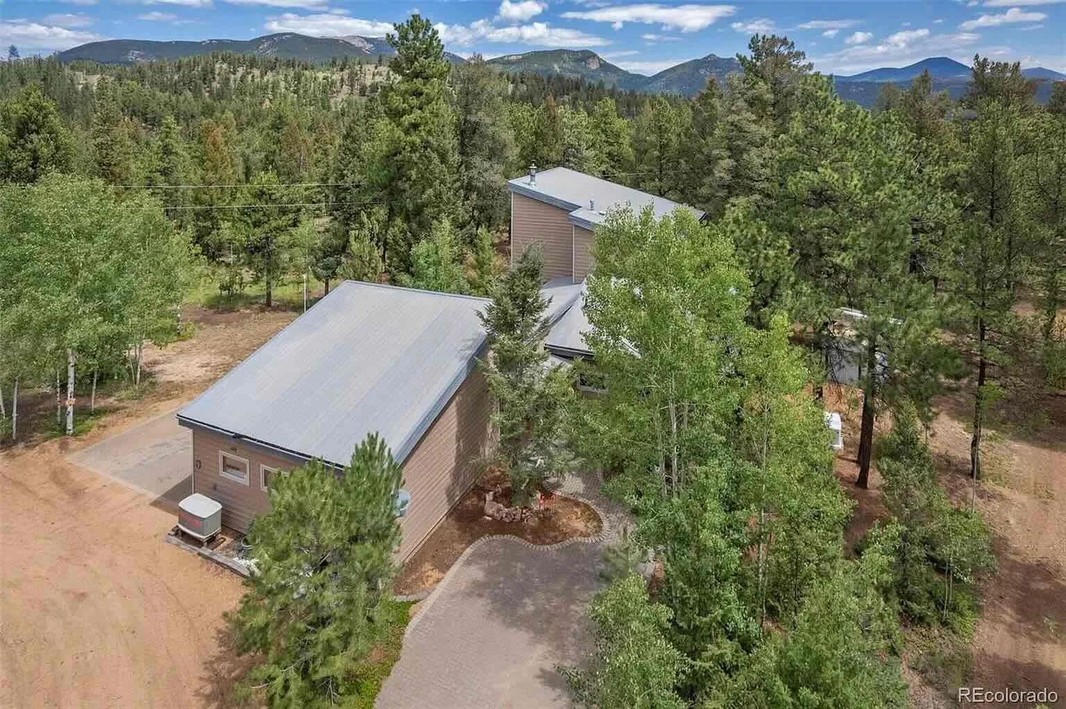 Aerial view of a house surrounded by trees with mountains in the background, clear blue sky, and a paved driveway.