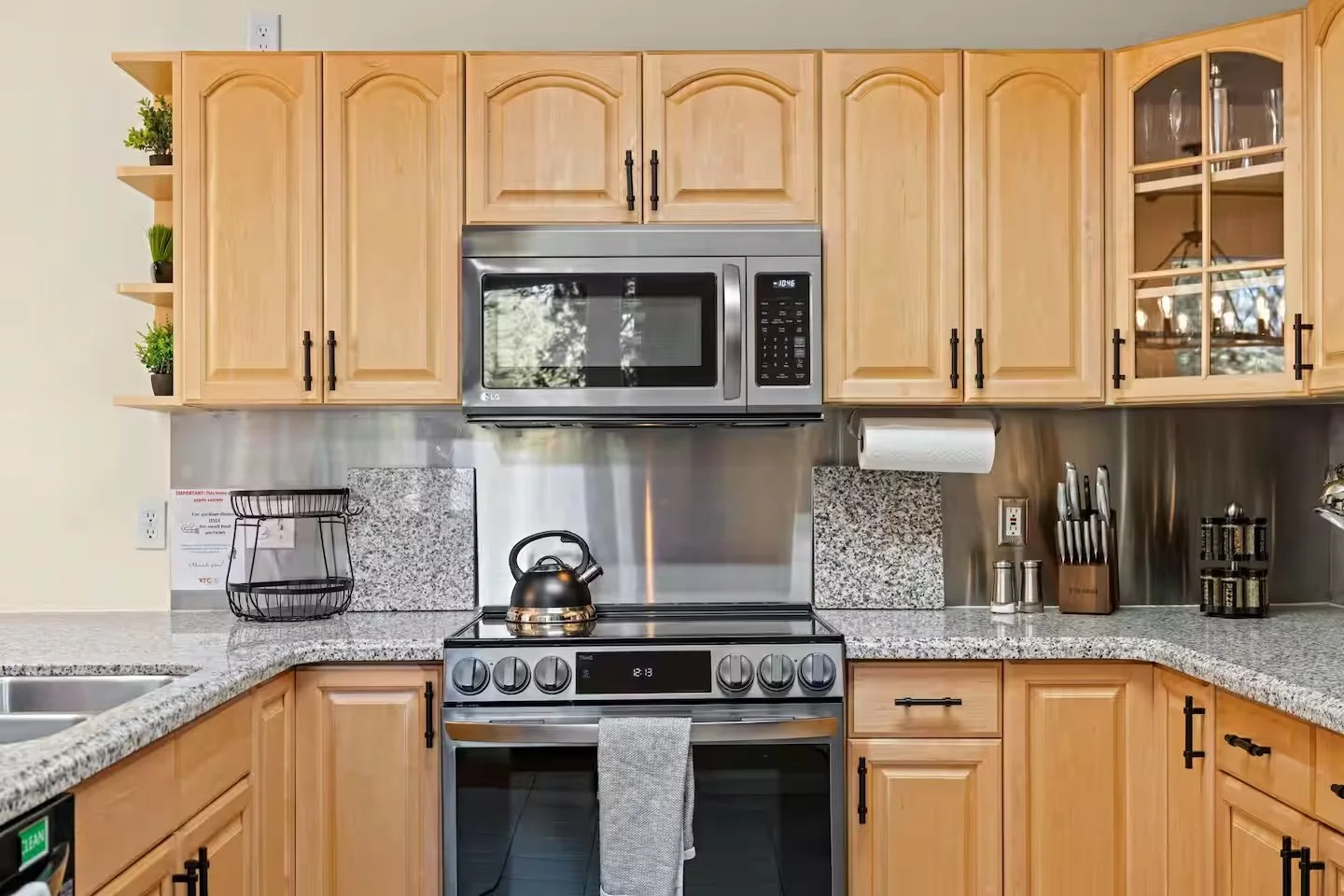 Kitchen with wooden cabinets, granite countertops, stainless steel oven and microwave, black kettle, knife set, spices, paper towel, and decorative plants.