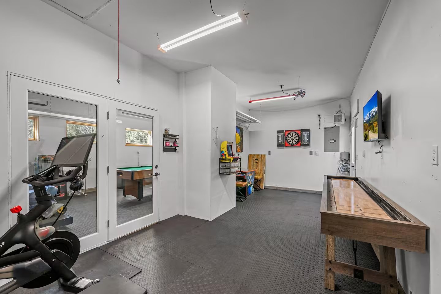 A recreational game room with arcade, dartboard, TV, and exercise bike.