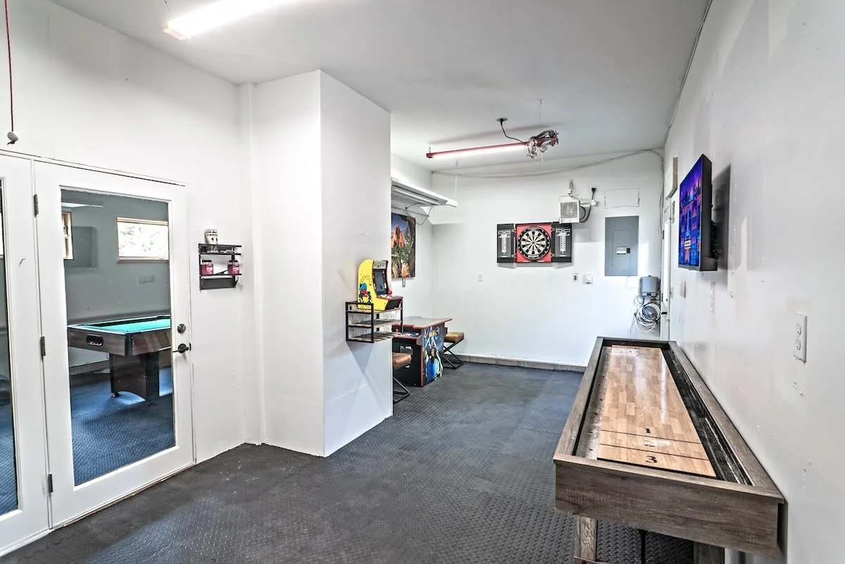 An indoor recreational room featuring a dartboard, a TV mounted on the wall, a shuffleboard table, and a small game area with an air hockey table visible through a glass door.